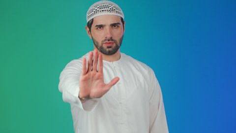 مفهوم الرفض والامتناع والانزعاج، الصمت والتوقف عن الكلام,بورتريه لشاب عربي خليجي اماراتي يرتدي الكندورة والطاقية يمد كفه نحو الامام  بتعابير النهي عن فعل شيء ما, ايماءات الحزم والجدية, خلفيه ملونة