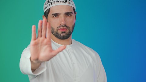 الصمت والتوقف عن الكلام, مفهوم الرفض والامتناع والانزعاج، بورتريه لشاب عربي خليجي اماراتي يرتدي الكندورة والطاقية يمد كفه نحو الامام  بتعابير النهي عن فعل شيء ما, ايماءات الحزم والجدية, خلفيه ملونة