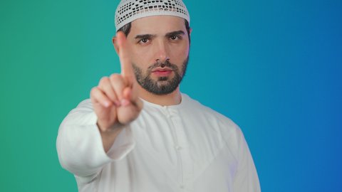 ايماءات الحزم والجدية,  مفهوم الرفض والامتناع والانزعاج، بورتريه لشاب عربي خليجي اماراتي يرتدي الكندورة والطاقية يرفع اصبع السبابة بتعابير النهي عن فعل شيء ما, خلفيه ملونة