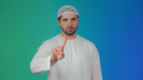 A portrait of a young Arab Gulf Emirati man wearing a kandura and a head cap, raising his index finger with expressions of disapproval of doing something, gestures of determination and seriousness, the concept of rejection, refusal, and discomfort, with a colorful background.