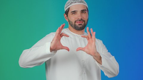 مفهوم المحبة والمودة، اظهار مشاعر الحب والشغف، بورتريه لشاب عربي خليجي اماراتي يرتدي الكندورة والطاقية يرفع يديه بإيماءات على شكل قلب، تعابير الفرح والسرور والسعادة، خلفية ملونة