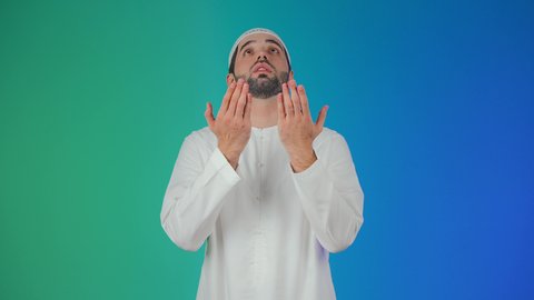 الطلب بترجي وتذلل من رب العباد, التضرع إلى الله تعالى, بورتريه لشاب عربي خليجي اماراتي مبتسم يرتدي الكندورة والطاقيه يرفع يديه بخشوع واستعطاف, طلب العون والرغبة في تحقيق الأمنيات, الوقوف بإيماءات الدعاء والتوسل والاسترحام,خلفية ملونة