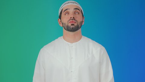 Standing with gestures of prayer, supplication, and seeking mercy, asking for help and the desire to fulfill wishes, pleading humbly and submissively to the Lord of the worlds, supplicating to God Almighty, a portrait of a smiling young Emirati Arab man wearing a kandura and a cap, raising his hands in reverence and pleading, with a colorful background.