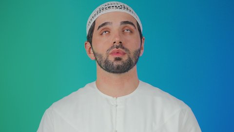 طلب العون والرغبة في تحقيق الأمنيات, الوقوف بإيماءات الدعاء والتوسل والاسترحام, الطلب بترجي وتذلل من رب العباد, التضرع إلى الله تعالى, بورتريه لشاب عربي خليجي اماراتي مبتسم يرتدي الكندورة والطاقيه يرفع يديه بخشوع واستعطاف, خلفية ملونة