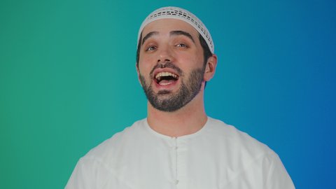 الشعور بالفرح والسعادة، ايماءات البهجة والسرور الشديد، بورتريه مقرب لرجل عربي اماراتي خليجي يرتدي الكندورة و الطاقيه ينظر الى الكاميرا بتعابير الضحك الهستيري, خلفية ملونه