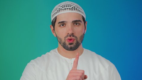 بورتريه لرجل عربي خليجي اماراتي يرتدي الكندورة و الطاقيه يضع أصبع السبابة على فمه بإيماءات طلب السكوت والصمت،التوقف عن الكلام وخفض الصوت، الهدوء والسكون وعدم الإزعاج، خلفية ملونه