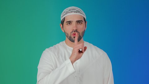 الهدوء والسكون وعدم الإزعاج، التوقف عن الكلام وخفض الصوت، بورتريه لرجل عربي خليجي اماراتي يرتدي الكندورة و الطاقيه يضع أصبع السبابة على فمه بإيماءات طلب السكوت والصمت،خلفية ملونه