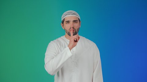 التوقف عن الكلام وخفض الصوت، بورتريه لرجل عربي خليجي اماراتي يرتدي الكندورة و الطاقيه يضع أصبع السبابة على فمه بإيماءات طلب السكوت والصمت، الهدوء والسكون وعدم الإزعاج، خلفية ملونه