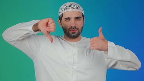 مفهوم الرفض والقبول،  الحيرة والتردد بخصوص امر ما، بورتريه لرجل عربي خليجي  اماراتي يرتدي الكندورة والطاقية يرفع اصبع الابهام للاعلى والاسفل بإيماءات عدم الموافقه والموافقه، عدم الإعجاب والإعجاب،  خلفية ملونه