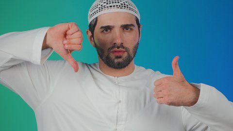 الحيرة والتردد بخصوص امر ما، بورتريه لرجل عربي خليجي  اماراتي يرتدي الكندورة والطاقية يرفع اصبع الابهام للاعلى والاسفل بإيماءات عدم الموافقه والموافقه، عدم الإعجاب والإعجاب، مفهوم الرفض والقبول،  خلفية ملونه