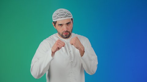 محاولة الدفاع عن النفس، احتراف رياضة الملاكمة، إغلاق اليدين والاستعداد للهجوم والمواجهة، مفهوم القوة والرجولة، بورتريه لرجل عربي خليجي اماراتي يرتدي الكندورة والطاقية ينظر للكاميرا بإيماءات الجدية والغضب،  خلفية ملونة