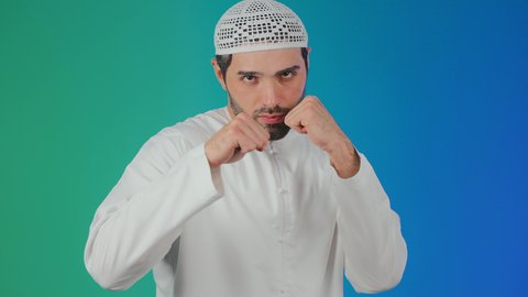 إغلاق اليدين والاستعداد للهجوم والمواجهة، احتراف رياضة الملاكمة،محاولة الدفاع عن النفس، مفهوم القوة والرجولة، بورتريه لرجل عربي خليجي اماراتي يرتدي الكندورة والطاقية ينظر للكاميرا بإيماءات الجدية والغضب،  خلفية ملونة