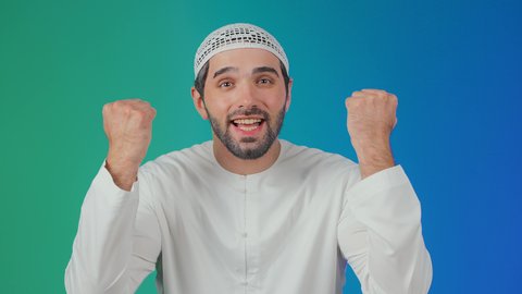 A portrait of a smiling Emirati Gulf Arab man wearing a kandura and a headscarf, raising his hands with expressions of interaction and enthusiasm, engaging in cheering for a football team, gestures of victory and triumph, colorful background.