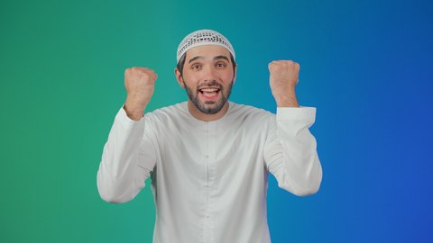 Gestures of victory and triumph, a portrait of a smiling Emirati Gulf Arab man wearing a kandura and a head cap, raising his hands with expressions of interaction and enthusiasm, engaging in cheering for the football team, colorful background.