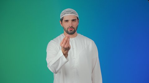 The concept of patience, deliberation, and caution, the threat and promise of something, a portrait of an Arab Gulf Emirati man wearing a kandura and a headscarf looking at the camera, raising his hand upwards with gestures asking for waiting, colorful background.