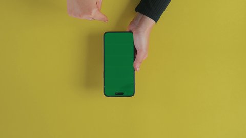 The concept of entertainment with modern technology and technology, fun and enjoyment during breaks, an overhead vertical shot of a hand of a Saudi Gulf Arab woman using a phone to browse social media, displaying an empty green chroma screen.