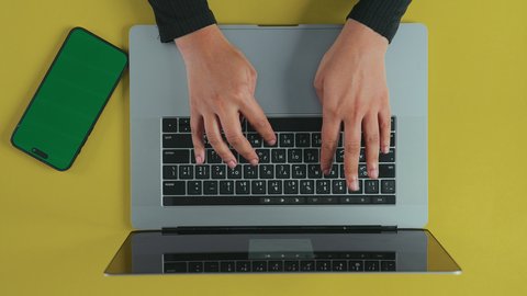 Sending an email via the computer, using modern and advanced technologies in work, holding meetings over the internet, an overhead vertical shot of the hand of an Arab Gulf Saudi woman wearing a black abaya using a laptop with a mobile phone beside her with a blank green screen, chroma.