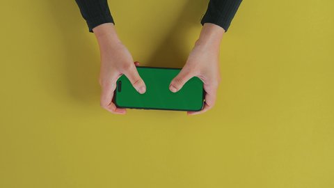 Using technology for entertainment, spending free time playing video games, browsing news on social media, an overhead vertical shot of the hand of an Arab Gulf Saudi woman wearing a black abaya and using a mobile phone, displaying a blank green screen on technical devices, chroma.