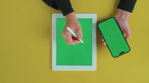 Using a pen to facilitate the explanation of work steps, monitoring social media, holding meetings online, an overhead vertical shot of the hand of an Arab Gulf Saudi woman wearing a black abaya using a tablet and holding a mobile phone with a blank green screen, chroma.