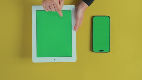 مفهوم العمل عن بعد،  متابعة مواقع التواصل الاجتماعي، تطبيقات وبرامج الأجهزة اللوحية، استخدام التكنولوجيا و التقنيات الحديثة، تصوير علوي عامودي ليد امرأة عربية خليجية سعودية ترتدي العباءة السوداء تستخدم التابلت و بجانبها هاتف محمول بشاشة خضراء فارغة, كروما