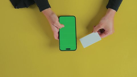 Online shopping, using modern payment methods, browsing social media platforms, displaying a blank green screen on technical devices, a vertical overhead shot of the hand of a Saudi Gulf Arab woman wearing a black abaya using a mobile phone and holding a credit card, chroma.