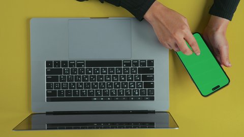 Comfortable office work environment, monitoring social media, holding meetings online, overhead vertical shot of the hand of an Arab Gulf Saudi woman wearing a black abaya using a laptop and holding a mobile phone with a blank green screen, chroma.