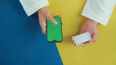 التسوق الالكتروني عبر الموبايل, استخدام وسائل الدفع الحديثة، تصفح مواقع التواصل الاجتماعي، عرض شاشة خضراء فارغة على الأجهزة التقنية، لقطة عمودية من الأعلى ليد رجل خليجي سعودي يرتدي الزي التقليدي يستخدم الجوال و يحمل بطاقة الائتمان, كروما