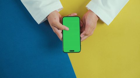 إنتظار مكالمة وإستخدام الميكروفون، عرض شاشة خضراء فارغة على الأجهزة التقنية، استخدام التكنولوجيا الحديثة لإنجاز المهام عن بُعد، لقطة عمودية من الأعلى ليد رجل خليجي سعودي يرتدي الزي التقليدي و يستخدم هاتفًا محمولًا, كروما