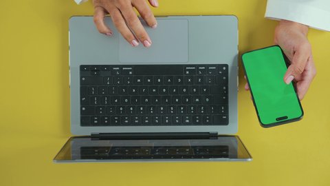 دور التكنولوجيا في في تسهيل الأعمال, الكتابة والنقر على الحاسوب محمول، متابعة مواقع التواصل الاجتماعي، عقد الاجتماعات عبر الانترنت، تصوير علوي عامودي ليد رجل عربي سعودي خليجي يرتدي الثوب التقليدي يستخدم اللابتوب و يحمل الهاتف المحمول بشاشة خضراء فارغة