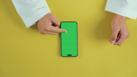 متابعة البرامج المفضلة، تصفح مواقع التواصل الاجتماعي، عرض شاشة خضراء فارغة على الأجهزة التقنية، استخدام التكنولوجيا الحديثة لإنجاز المهام عن بُعد، لقطة عمودية من الأعلى ليد رجل خليجي سعودي يرتدي الزي التقليدي وهو يستخدم هاتفًا محمولًا, كروما