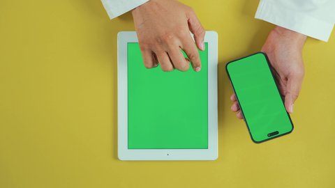 Display a blank green screen on technical devices, using modern technology to accomplish tasks remotely, an overhead vertical shot of the hand of an Arab Saudi Gulf man wearing traditional attire using a tablet and holding a mobile phone, browsing social media sites, chroma.
