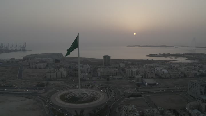 راية التوحيد الخضراء ترفر في السماء، كورنيش جدة البحري، ميدان وشوارع ومعالم جدة المشهورة، تصوير درون لطريق الملك عبد الله المالي في مدينة جدة في المملكة العربية السعودية في وقت الغروب، حركة مرور السيارات، الابراج والمباني السكنية، جمال مدينة جدة