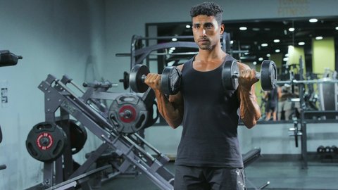 Strength, challenge, and motivation for a healthy lifestyle, physical activity to maintain health and physical fitness, lifting heavy weights, strengthening and building body muscles, weightlifting exercises, a young Arab Gulf Saudi man wearing sportswear practicing exercises with dumbbells.