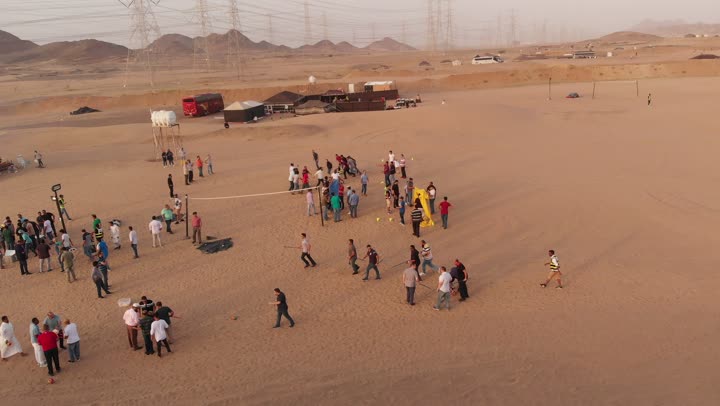 Adventure and exploration, the landscapes and soft golden sands in the desert, the soft golden dunes and natural terrain in the Kingdom of Saudi Arabia, the concept of mobility and movement, aerial drone footage of a group of Arab Saudi Gulf individuals walking somewhere in the desert.