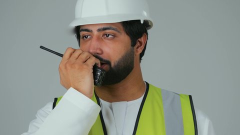 قيادة العمل من خلال إصدار الأوامر، متابعة سير الاعمال، العمل في القطاع الهندسي،  بورتريه مقرب لمهندس عربي خليجي إماراتي يرتدي الكندورة والخوذة وسترة الحماية يقوم باستخدام جهاز الاتصال اللاسلكي، مهن وظائف هندسية، خلفية بيضاء