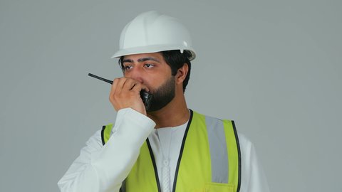 Issuing orders to carry out tasks, monitoring the progress of work, working in the engineering sector, a portrait of an Emirati Gulf Arab engineer wearing a kandura, helmet, and safety vest using a wireless communication device, engineering job professions, white background.