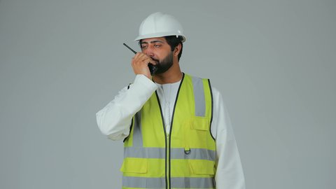 منح التعليمات لتنفيذ العمل، متابعة سير الاعمال، العمل في القطاع الهندسي،  بورتريه لمهندس عربي خليجي إماراتي يرتدي الكندورة والخوذة وسترة الحماية يقوم باستخدام جهاز الاتصال اللاسلكي، مهن وظائف هندسية، خلفية بيضاء