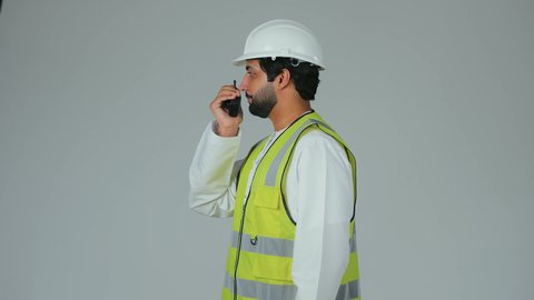 تحديد المهام وتوجيه الأوامر، متابعة سير الاعمال، العمل في القطاع الهندسي،  بورتريه من الجانب لمهندس عربي خليجي إماراتي يرتدي الكندورة والخوذة وسترة الحماية يقوم باستخدام جهاز الاتصال اللاسلكي، مهن وظائف هندسية، خلفية بيضاء