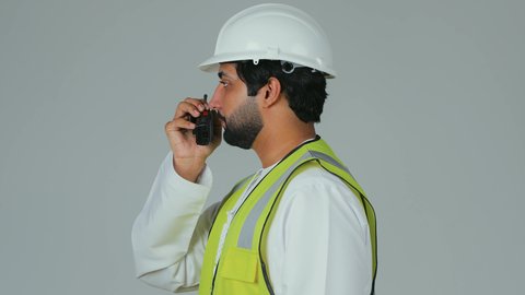 إعطاء أوامر محددة للموظفين، متابعة سير الاعمال، العمل في القطاع الهندسي،  بورتريه مقرب من الجانب لمهندس عربي خليجي إماراتي يرتدي الكندورة والخوذة وسترة الحماية يقوم باستخدام جهاز الاتصال اللاسلكي، مهن وظائف هندسية، خلفية بيضاء