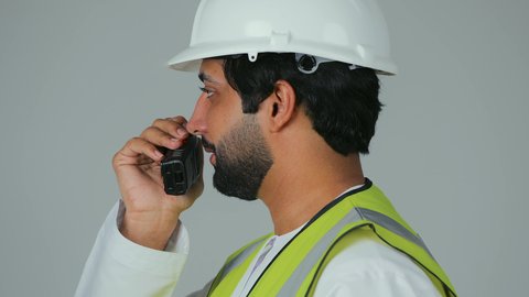 توجيه الفريق لتنفيذ المهام، متابعة سير الاعمال، العمل في القطاع الهندسي،  بورتريه مقرب من الجانب لمهندس عربي خليجي إماراتي يرتدي الكندورة والخوذة وسترة الحماية يقوم باستخدام جهاز الاتصال اللاسلكي، مهن وظائف هندسية، خلفية بيضاء
