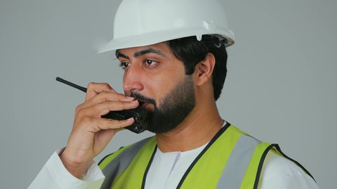 إصدار التعليمات الخاصة بالمشاريع، متابعة سير الاعمال، العمل في القطاع الهندسي،  بورتريه مقرب لمهندس عربي خليجي إماراتي يرتدي الكندورة والخوذة وسترة الحماية يقوم باستخدام جهاز الاتصال اللاسلكي، مهن وظائف هندسية، خلفية بيضاء