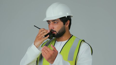 إدارة العمل من خلال إعطاء الأوامر، متابعة سير الاعمال، العمل في القطاع الهندسي، الإشراف على المشاريع،  بورتريه لمهندس عربي خليجي إماراتي يرتدي الكندورة والخوذة وسترة الحماية يقوم باستخدام جهاز الاتصال اللاسلكي، مهن وظائف هندسية، خلفية بيضاء