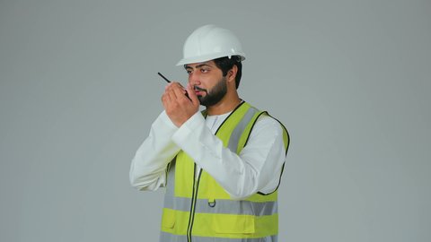 إصدار توجيهات مرتبطة بالعمل، متابعة سير الاعمال، العمل في القطاع الهندسي، الإشراف على المشاريع،  بورتريه لمهندس عربي خليجي إماراتي يرتدي الكندورة والخوذة وسترة الحماية يقوم باستخدام جهاز الاتصال اللاسلكي، مهن وظائف هندسية، خلفية بيضاء