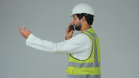 Giving instructions for task performance, monitoring the progress of work, working in the engineering sector, supervising projects, a side portrait of an Emirati Gulf Arab engineer wearing a kandura, helmet, and safety vest using a wireless communication device, engineering job professions, white background.