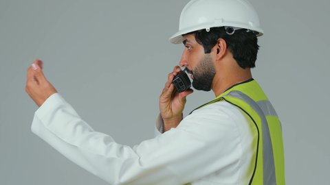 Issuing task orders, monitoring the progress of work, working in the engineering sector, supervising projects, a close-up side portrait of an Emirati Gulf Arab engineer wearing a kandura, helmet, and safety vest using a wireless communication device, engineering job professions, white background.