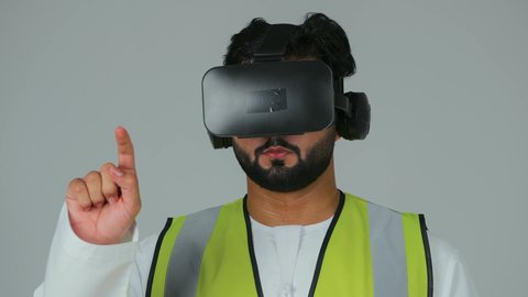 The use of virtual engineering in an interactive way, embodying and viewing engineering projects relying on technology, a close-up portrait of an Emirati Gulf Arab engineer wearing a kandura and work jacket and virtual reality glasses, raising his hands to touch holographic elements, white background.