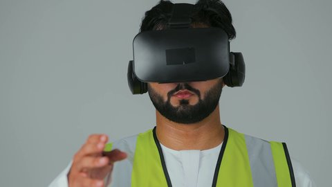 الاندماج مع الهندسة الافتراضية، بورتريه مقرب لمهندس عربي خليجي إماراتي يرتدي الكندورة والسترة الخاصة بالعمل ونظارة الواقع الافتراضي يرفع يديه لمحاولة تحجيم العناصر الهولوجرامية والإمساك بها، تجسيد ومشاهدة المشاريع الهندسية بالاعتماد على التكنولوجيا، خلفية بيضاء