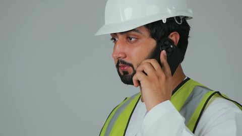 Remote work and tracking achievements, using modern technological devices, youth professions and jobs, working in the engineering sector, a close-up portrait of an Emirati Gulf Arab engineer wearing a kandura, helmet, and safety vest, holding a mobile phone and making a phone call, integrating advanced technology with engineering, white background.