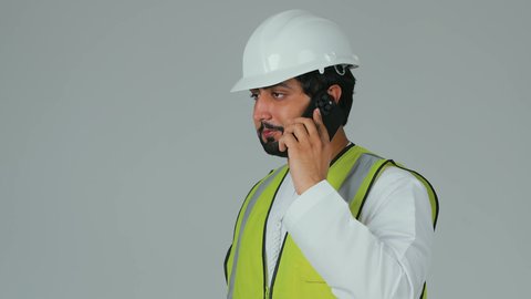 Remote task management, using modern technological devices, youth professions and jobs, working in the engineering sector, a portrait of an Emirati Gulf Arab engineer wearing a kandura, helmet, and safety vest holding a mobile phone making a phone call, integrating advanced technology with engineering, white background.