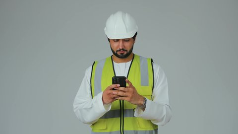 Integrating advanced technology into work, using technical devices to supervise work, the concept of architecture, a portrait of an Arab Gulf Emirati engineer wearing a kandura, helmet, and safety vest holding a mobile phone in his hand, white background.